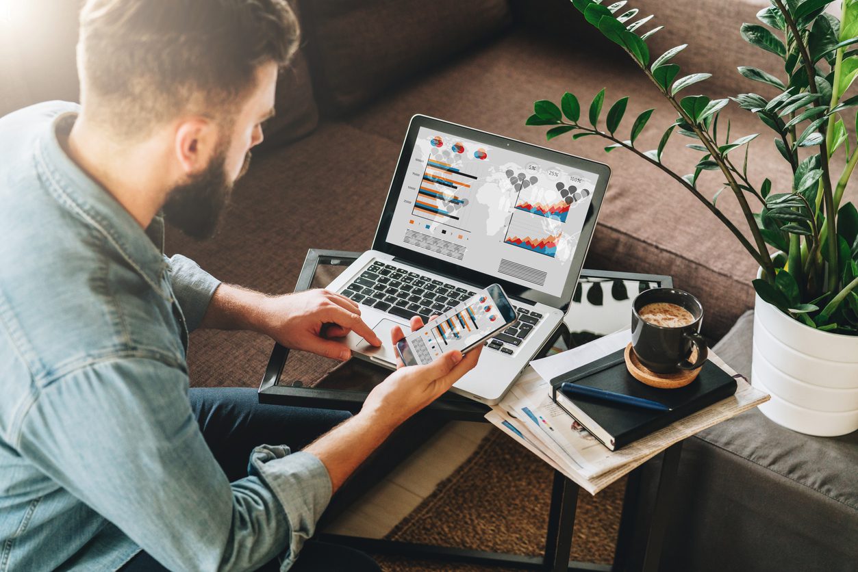 man-entrepreneur-freelancer-sits-at-home-on-couch-at-coffee-table-uses-smartphone-working-on-laptop-with-graphs